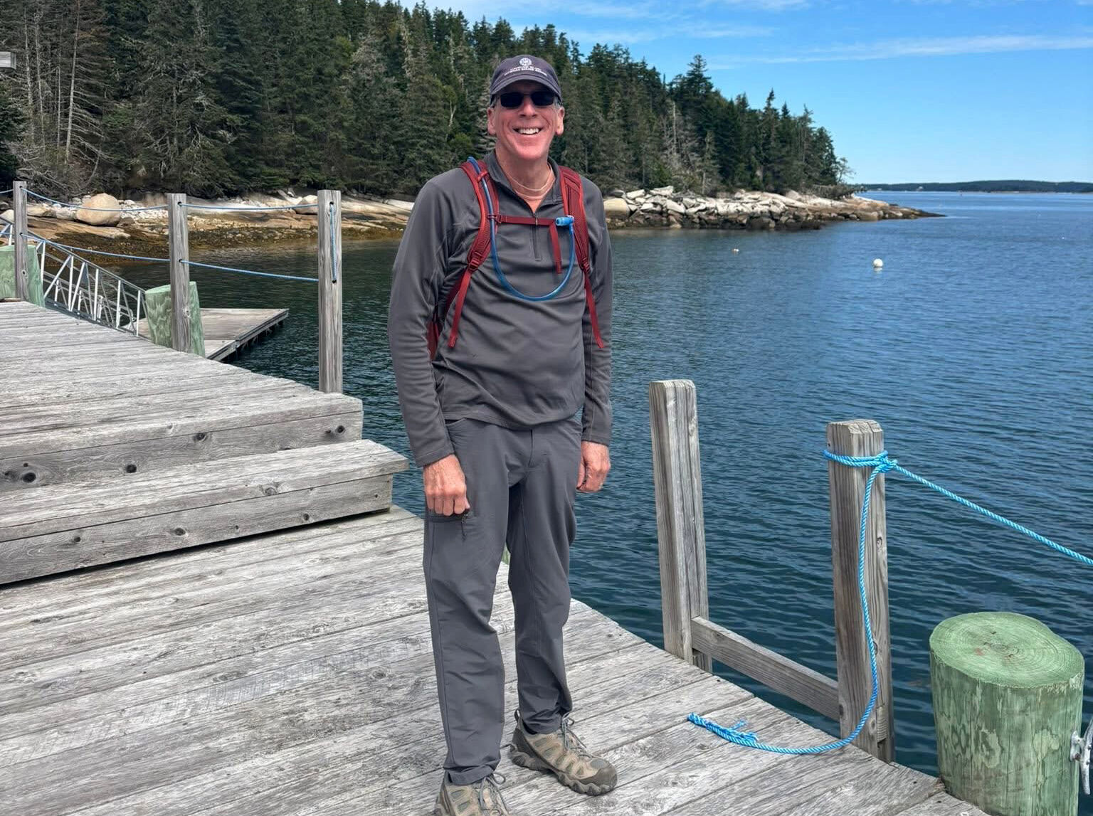 The image shows a man standing on a wooden dock. He is wearing a gray long-sleeved shirt, gray pants, a backpack, and a cap. The background features a body of water, trees, and a clear sky. The man is smiling, and the overall scene suggests a sunny day outdoors.
